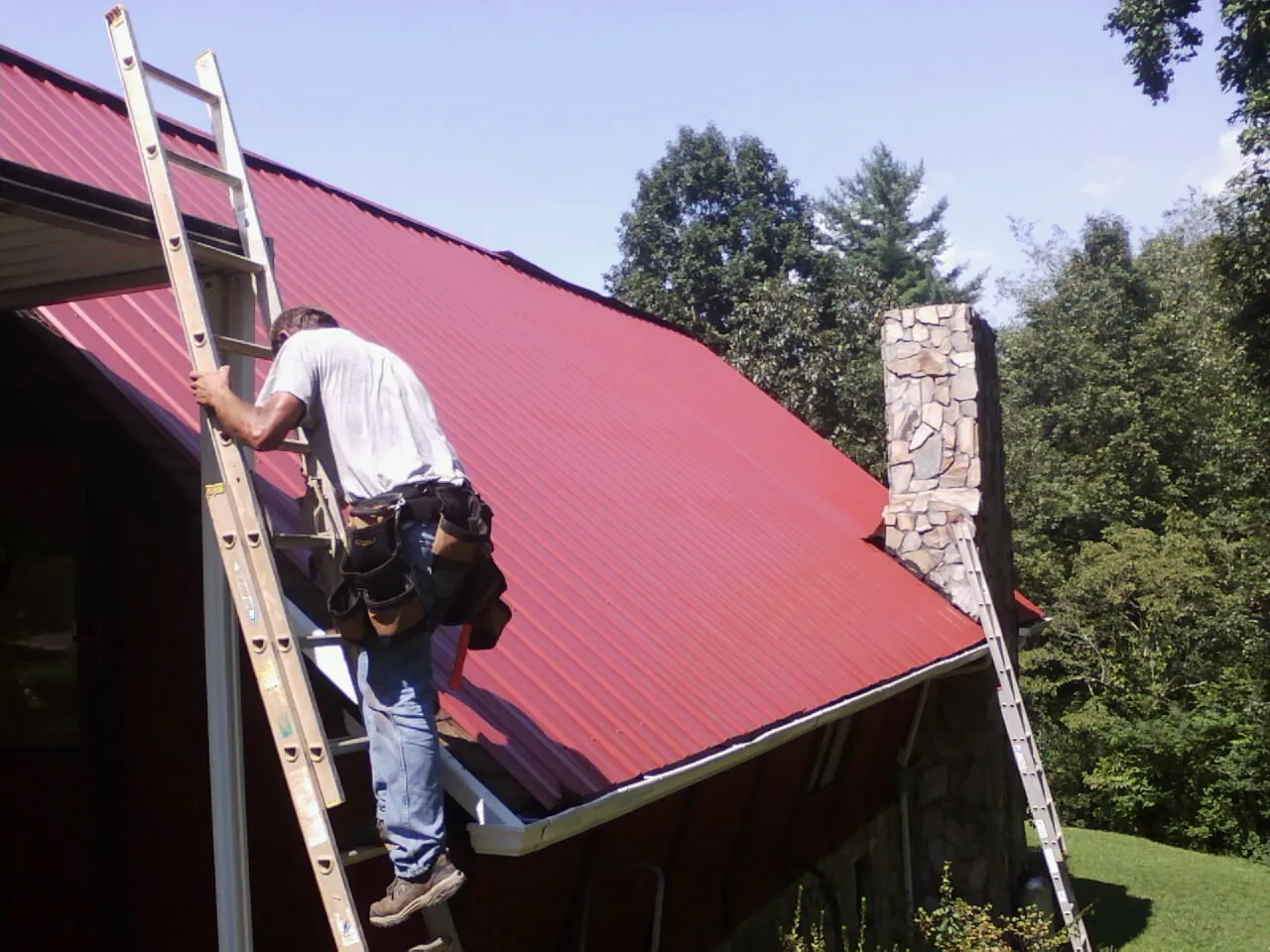 Roofer on a ladder beside a red metal roof and stone chimney — Soffit & Fascia Repair by Roofing Brothers in Plymouth